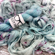 Load image into Gallery viewer, A skein of light teal blue merino linen yarn with  speckles of pink and purple a label that reads 'Feisty Fibres', surrounded by unspooled yarn.