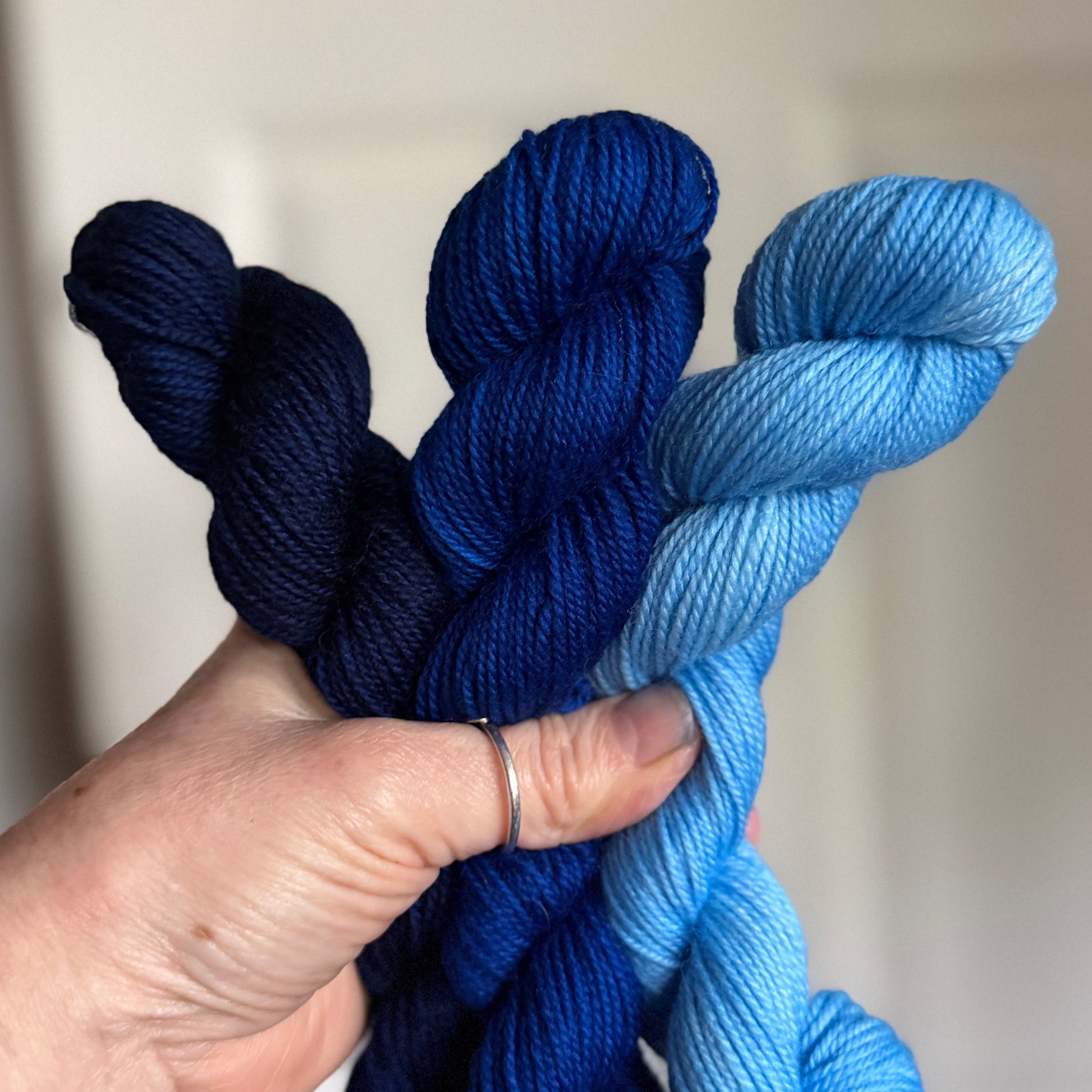 Three skeins of yarn held in a hand, showing color gradients from dark navy to medium blue and light sky blue, against a softly blurred indoor background.